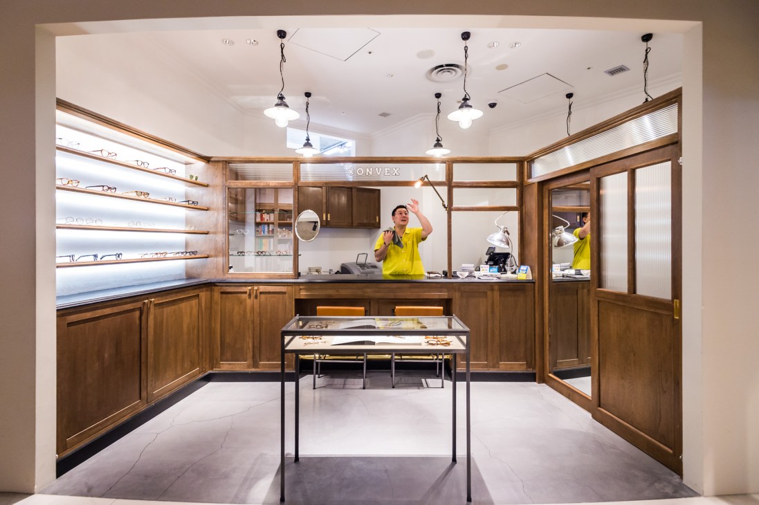 Convex vintage eyewear shop interior with wood cabinetry, illuminated shelving displaying glasses, and a shopkeeper in yellow shirt at the counter.