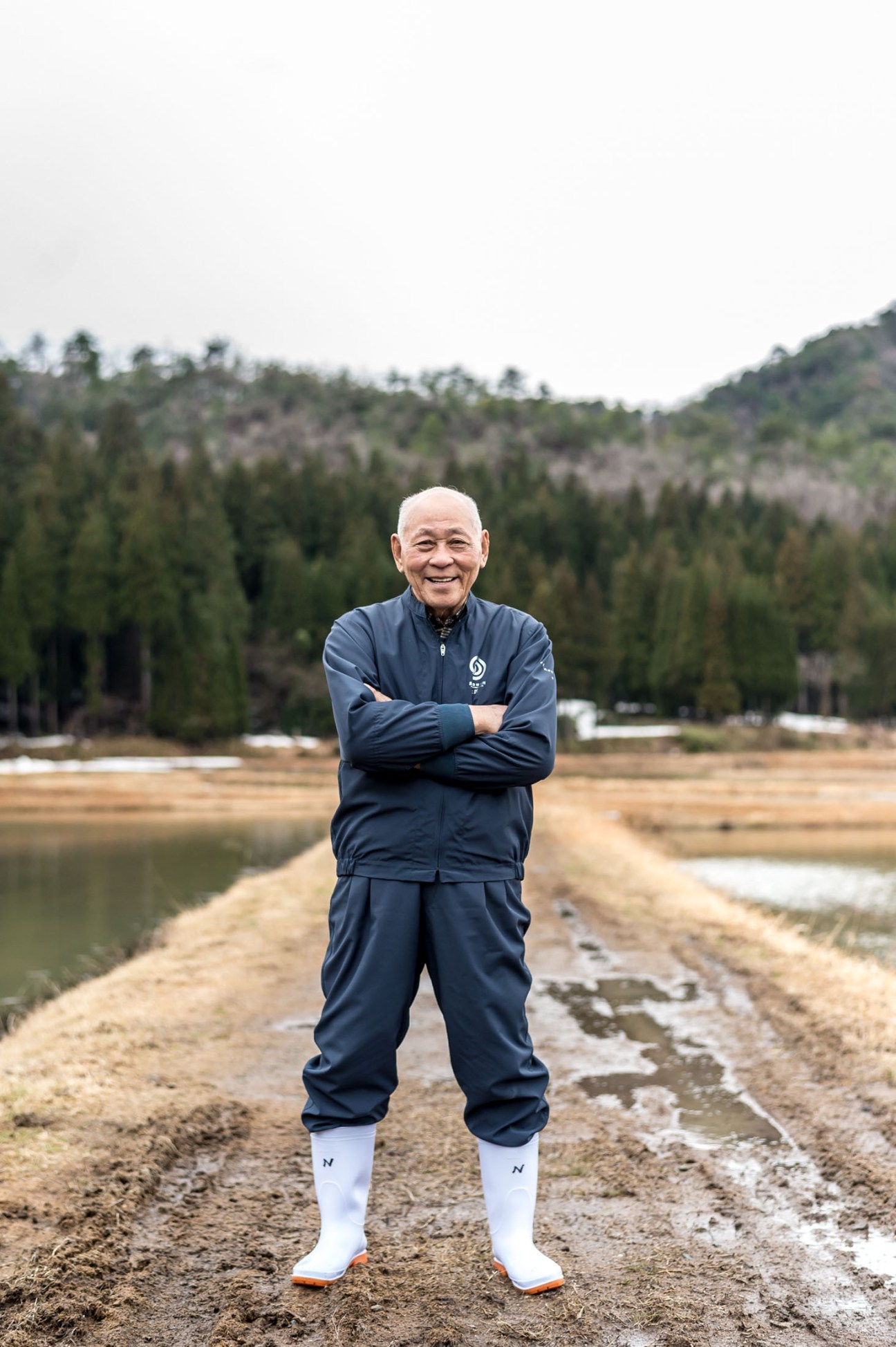 Naohiko Noguchi, an elderly Japanese man in blue athletic wear, stands smiling with arms crossed in a mountain valley with forested hills behind him.