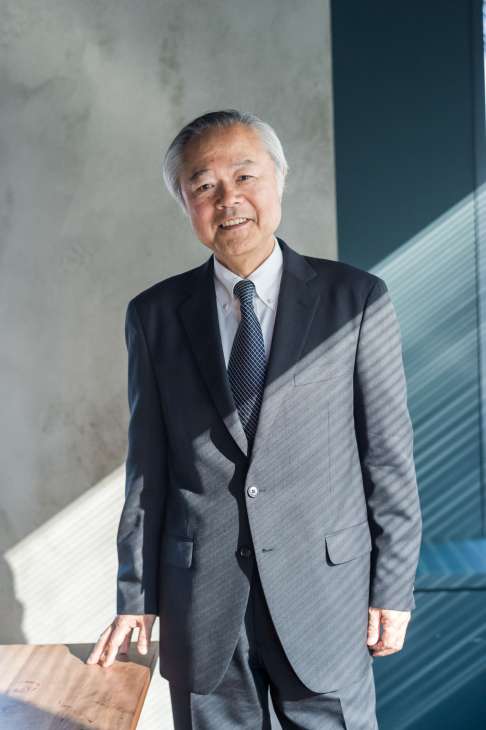Setouchi Cruise president Akio Jo stands in a grey suit and striped tie in front of a concrete wall and teal panel.