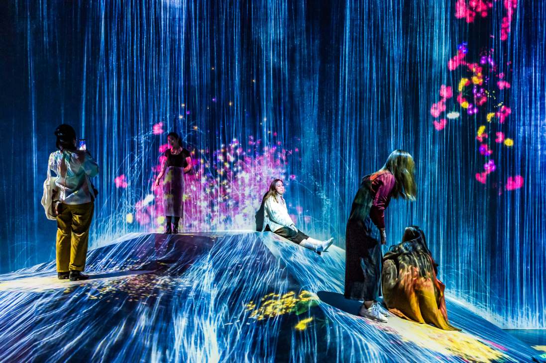 Visitors interact with TeamLab's 'Universe of Water Particles on a Rock Where People Gather' digital art installation with blue streaks and colorful floating particles.
