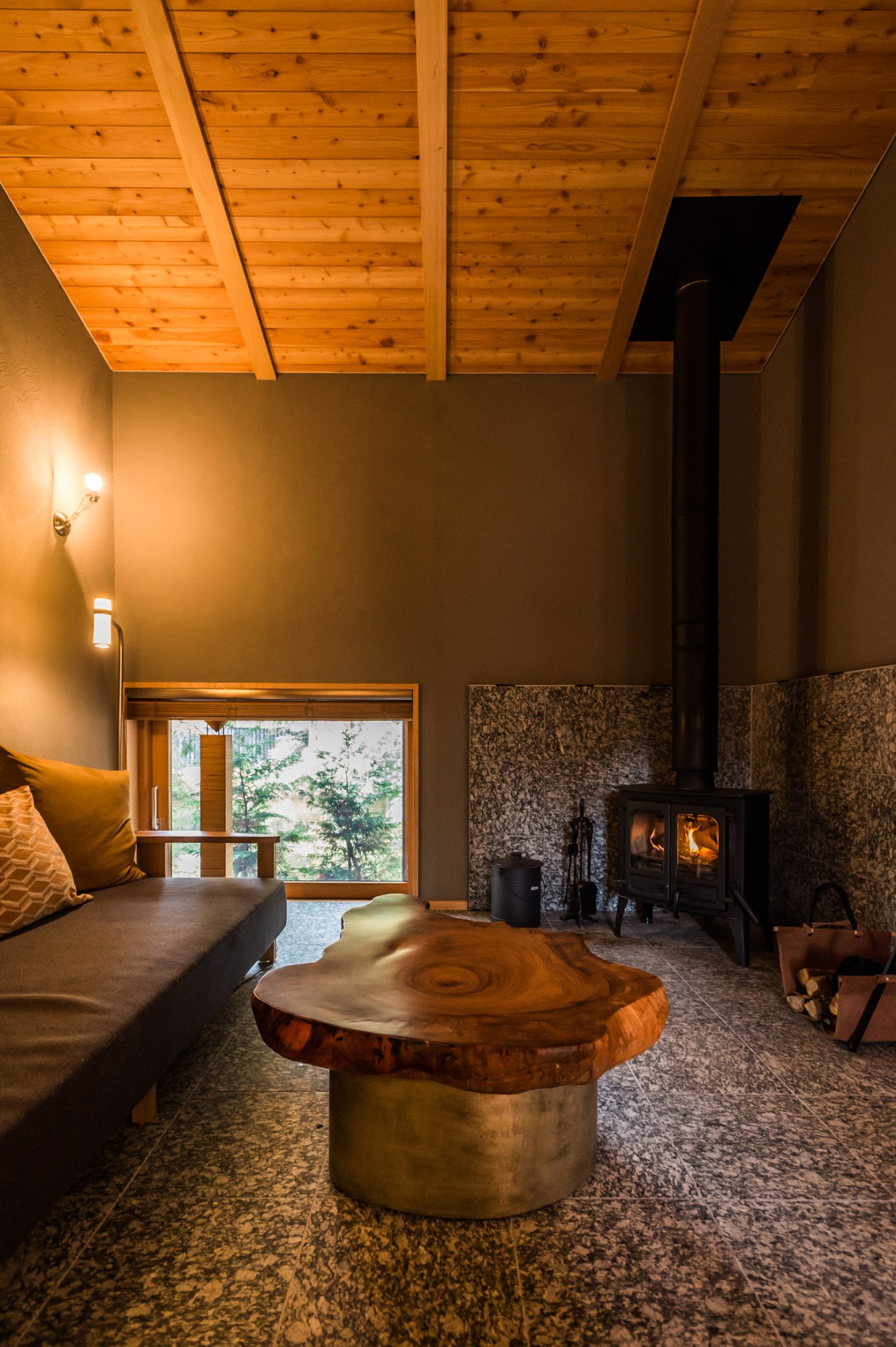 Modern villa interior with wooden ceiling, wood-slice table, fireplace, and large window overlooking forest.