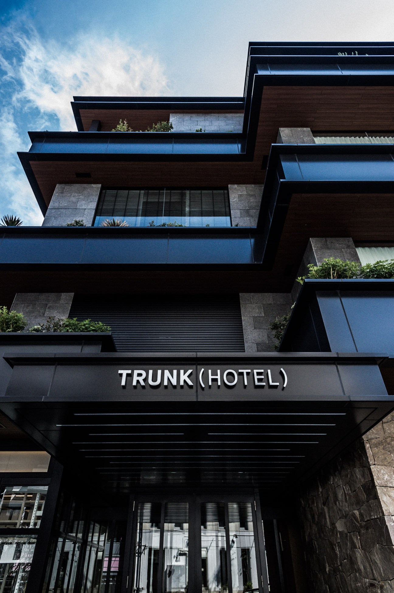 Trunk Hotel's modern facade featuring dark wood and blue glass panels with geometric overhanging balconies against a blue sky.