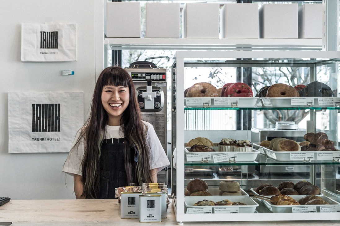 Woman smiling behind counter at Trunk Hotel shop with display case of donuts and baked goods.