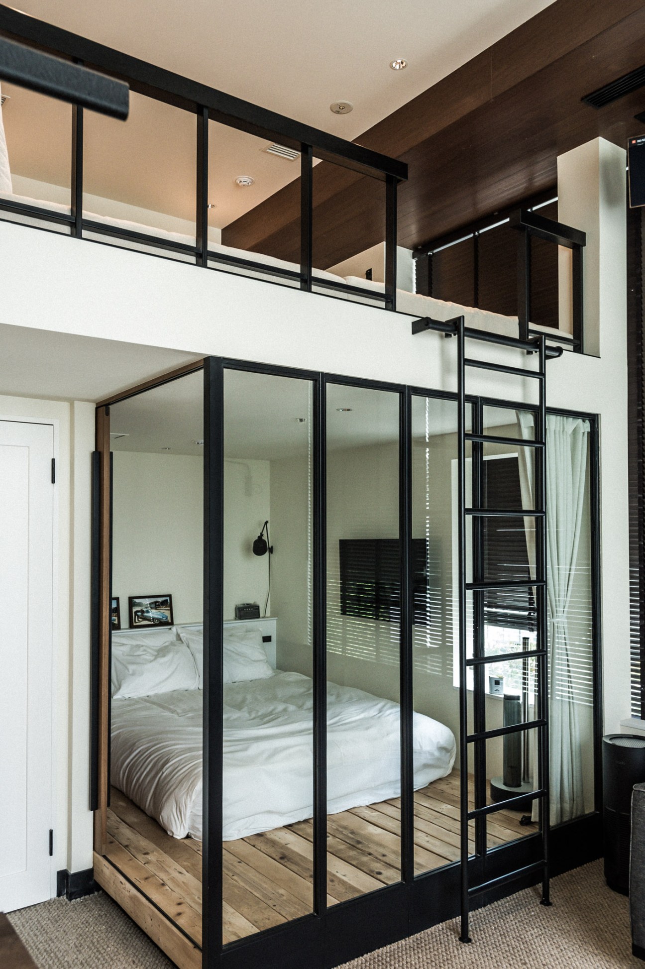 Modern hotel bedroom with glass-enclosed bed frame, black metal framework, white bedding, and mezzanine level above.