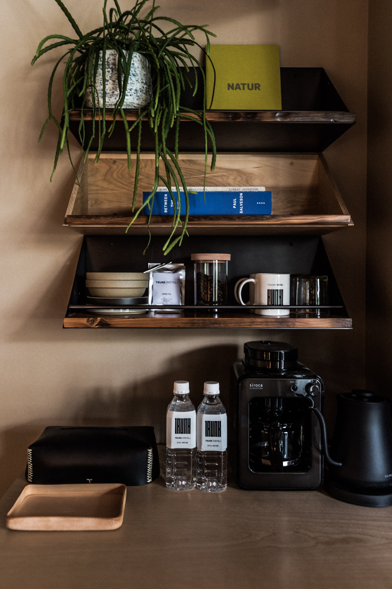 Trunk Hotel in-room amenities displayed on wooden shelves including books, a potted plant, coffee maker, water bottles and toiletries.