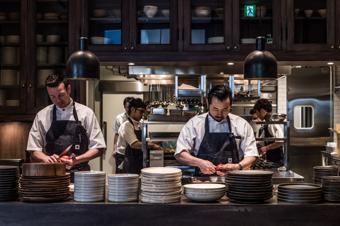 Chefs in white shirts and dark aprons prepare food in a modern kitchen with stacked plates and hanging pendant lights.
