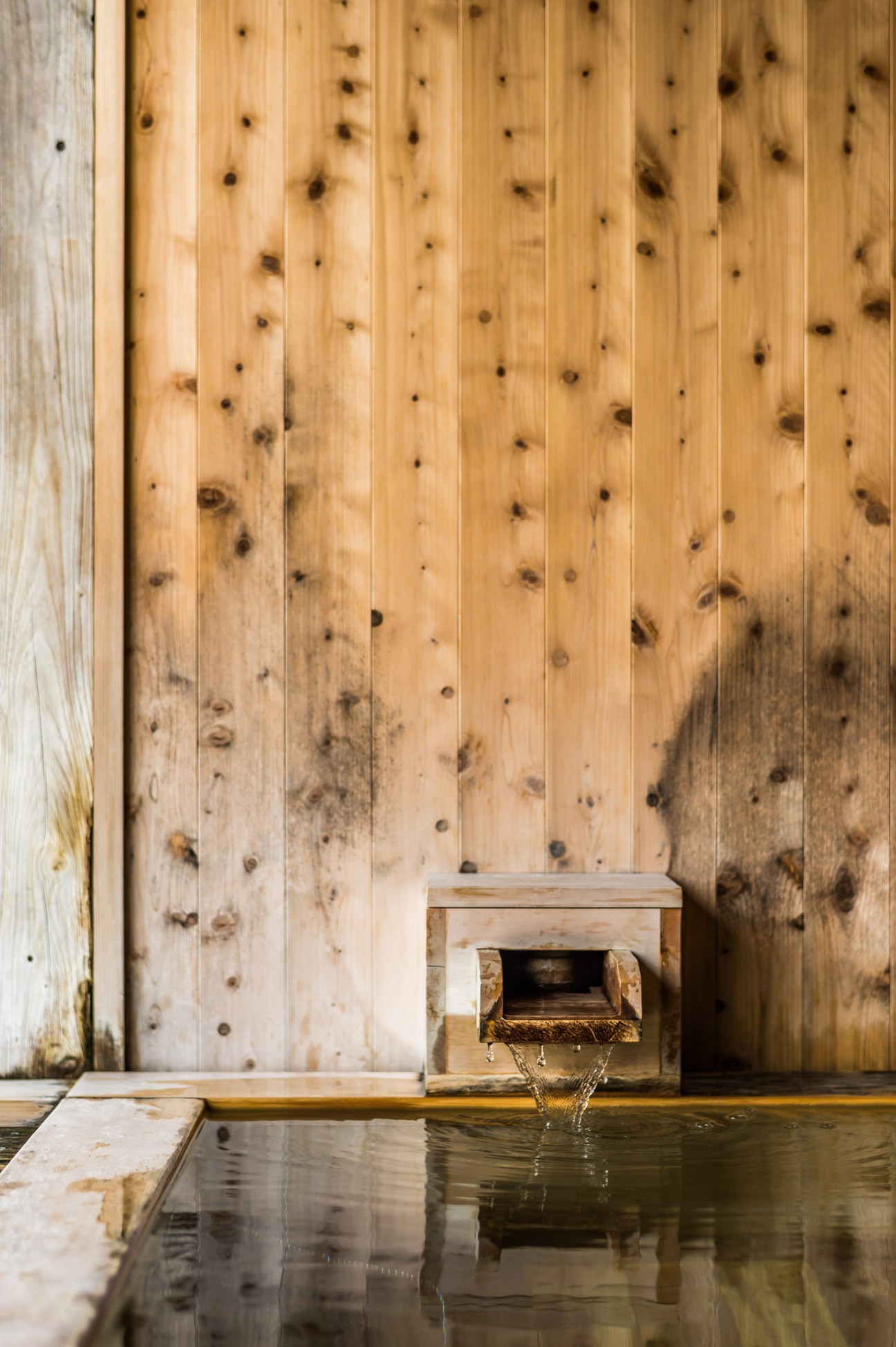 Natural hot spring water flowing from a wooden spout into a dark water basin against hinoki cypress wood paneling.