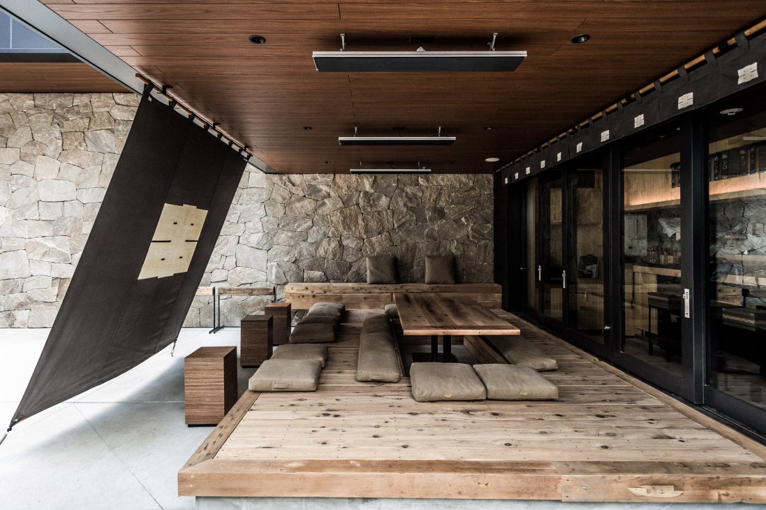 Modern lounge area with wooden platform, cushioned seating, stone wall, and black-framed glass doors at Trunk Hotel.