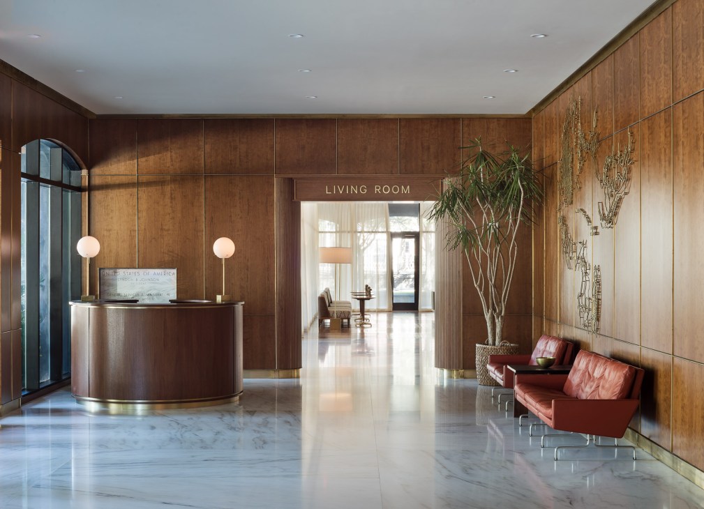Luxury hotel lobby with wood-paneled walls, circular reception desk, pendant lights, red leather seating and potted plant.