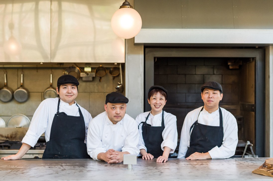 Four restaurant staff members in white chef coats and black aprons stand at a counter in a modern kitchen.