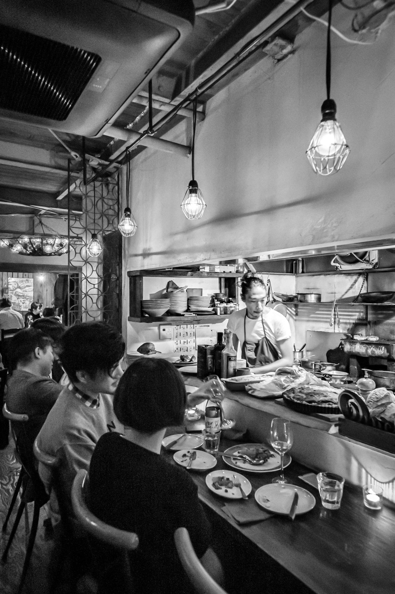Black and white photo of diners at counter watching chef prepare food in open kitchen restaurant