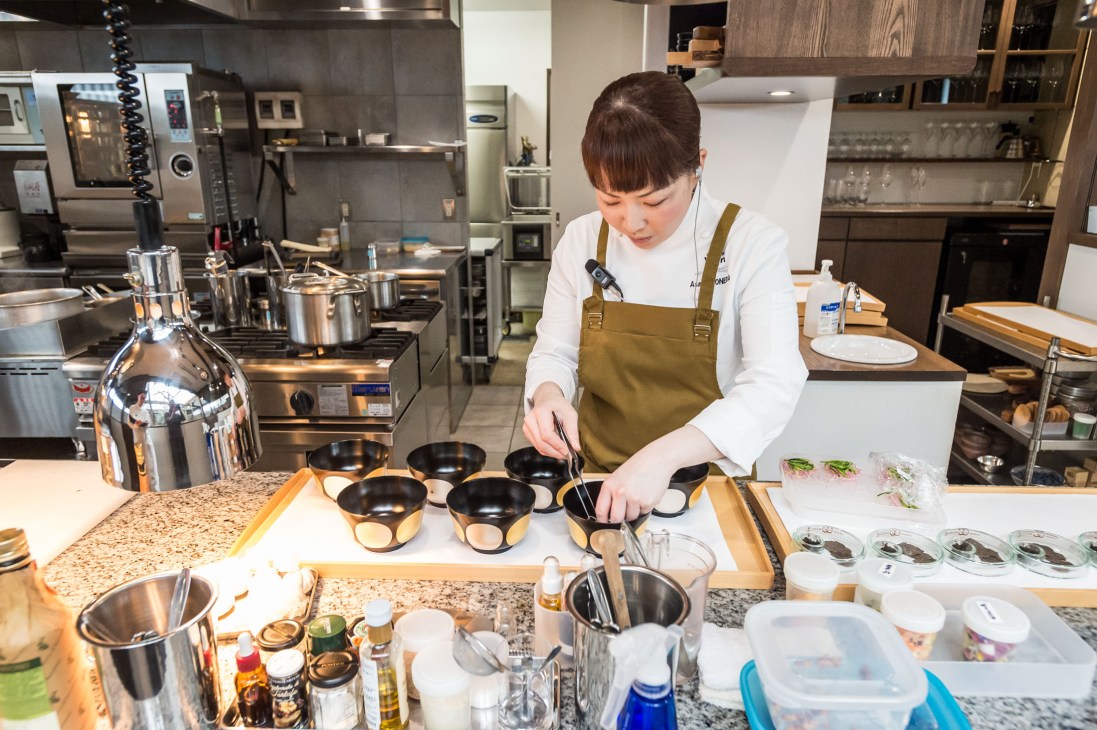 A chef in a mustard apron arranges black and gold bowls on a granite counter in a modern kitchen at Shokudo Yarn.