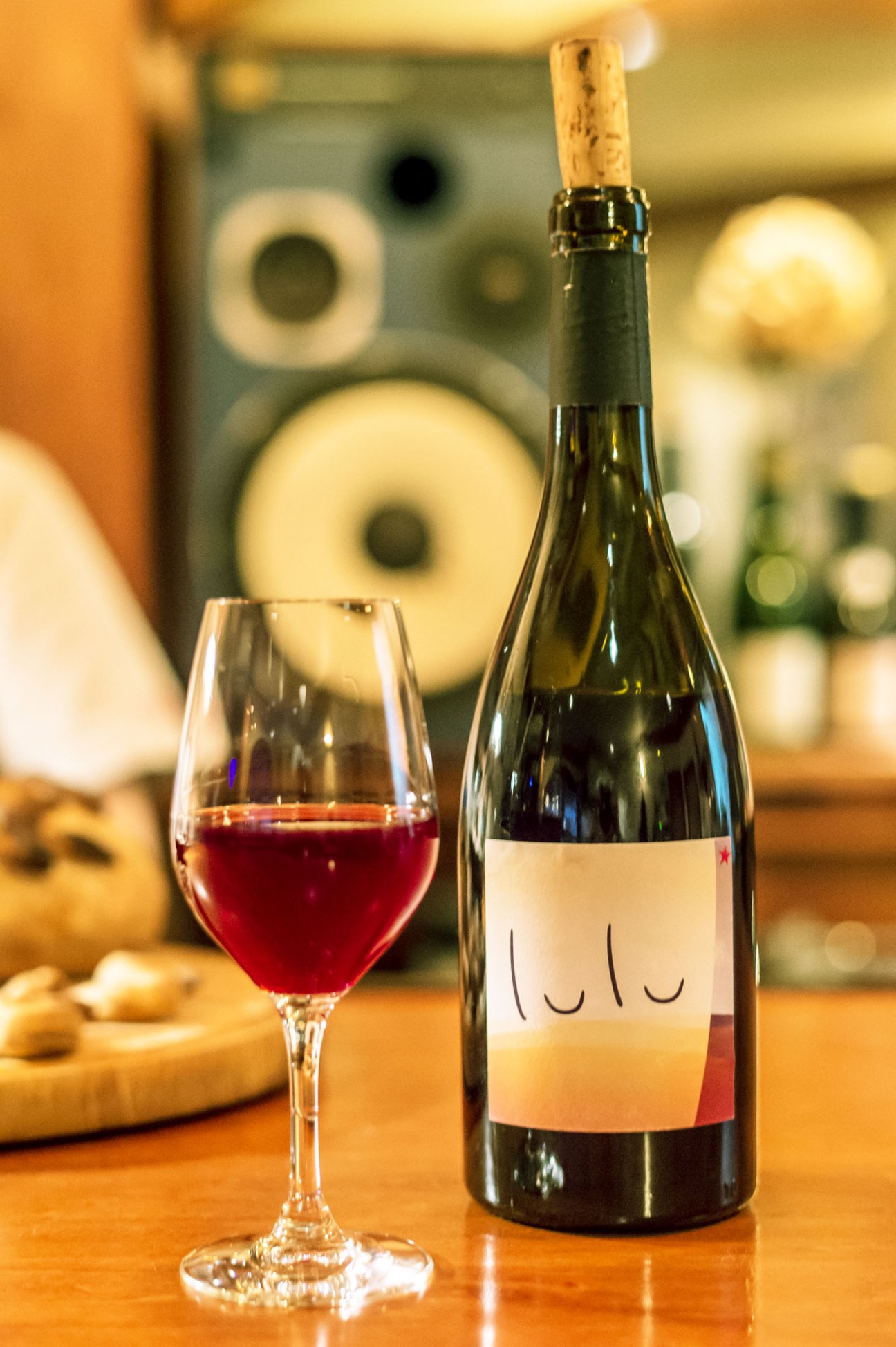 Red wine in a glass next to a bottle labeled 'lulu' with speakers blurred in background.