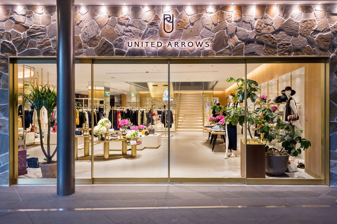 United Arrows storefront with glass facade showing interior displays and stone wall with illuminated brand signage