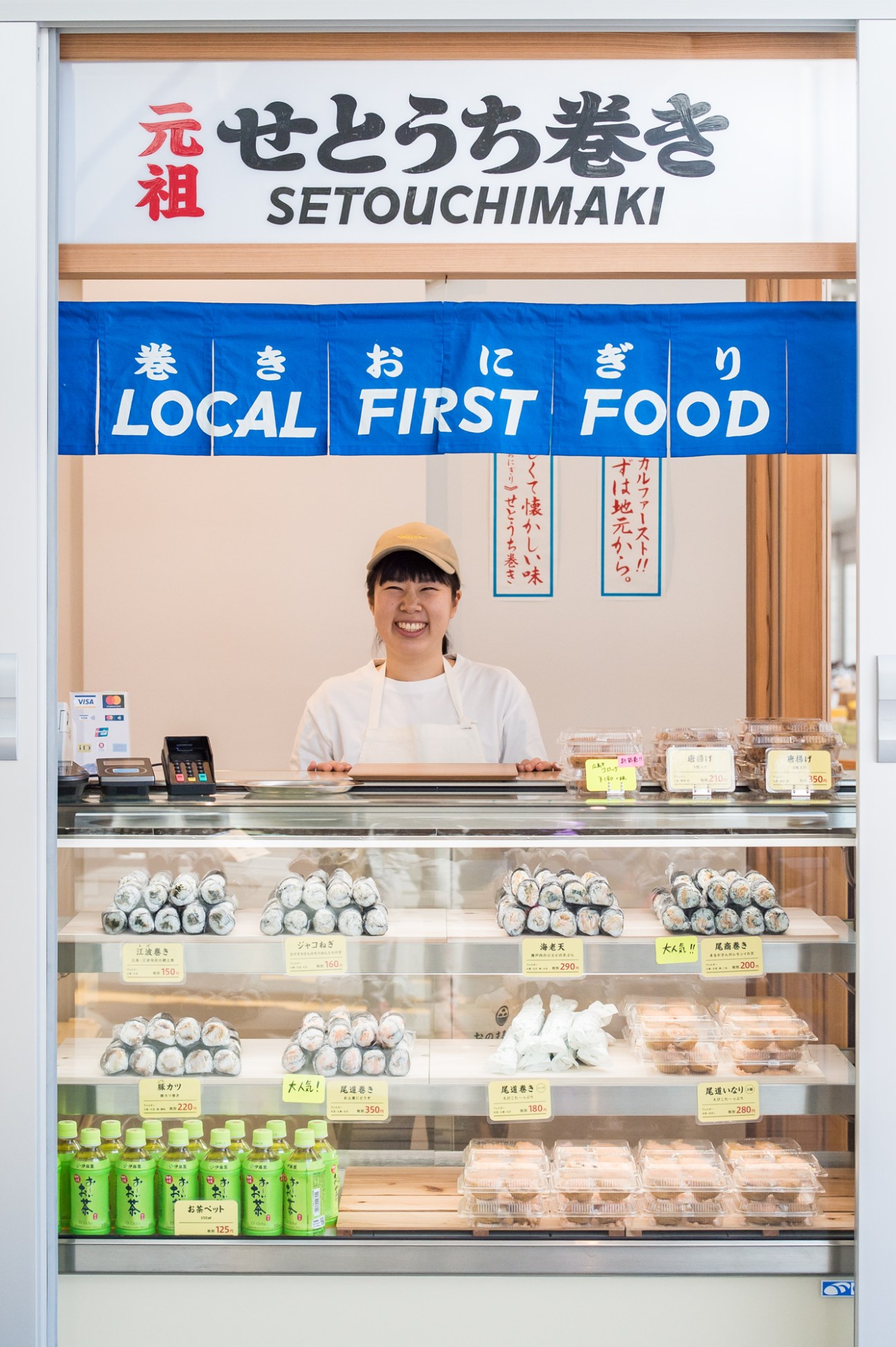 Staff member at a kiosk counter displaying setouchi-maki sushi rolls and other local food items for sale.
