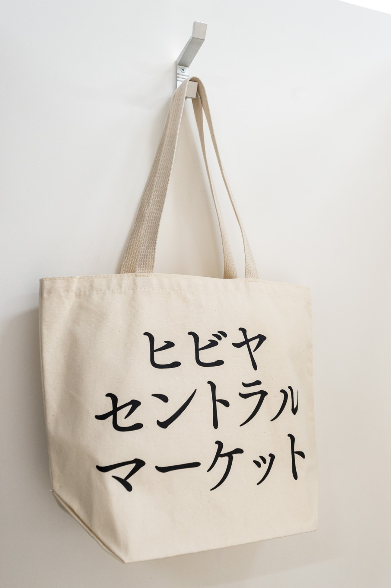 Cream-colored canvas tote bag with Japanese characters printed in black, hanging from a metal hook.