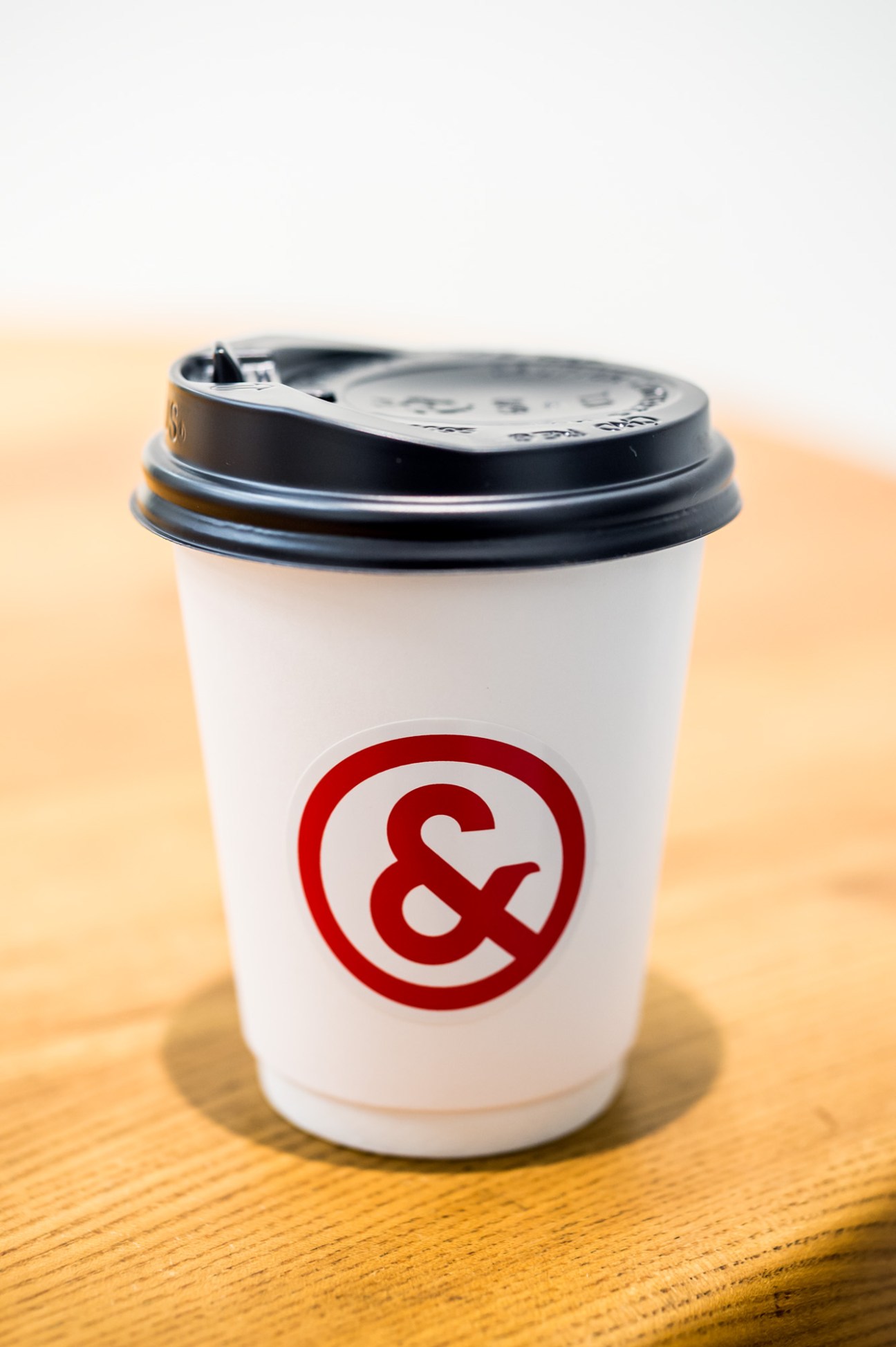 A white coffee cup with black lid branded with And Coffee Roasters' red ampersand logo on wooden surface.