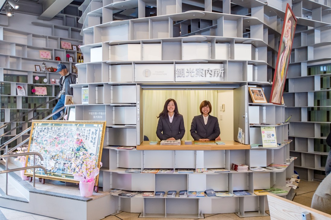 Two women staff members at the Onomichi Station tourist information desk with shelving and local maps displayed.
