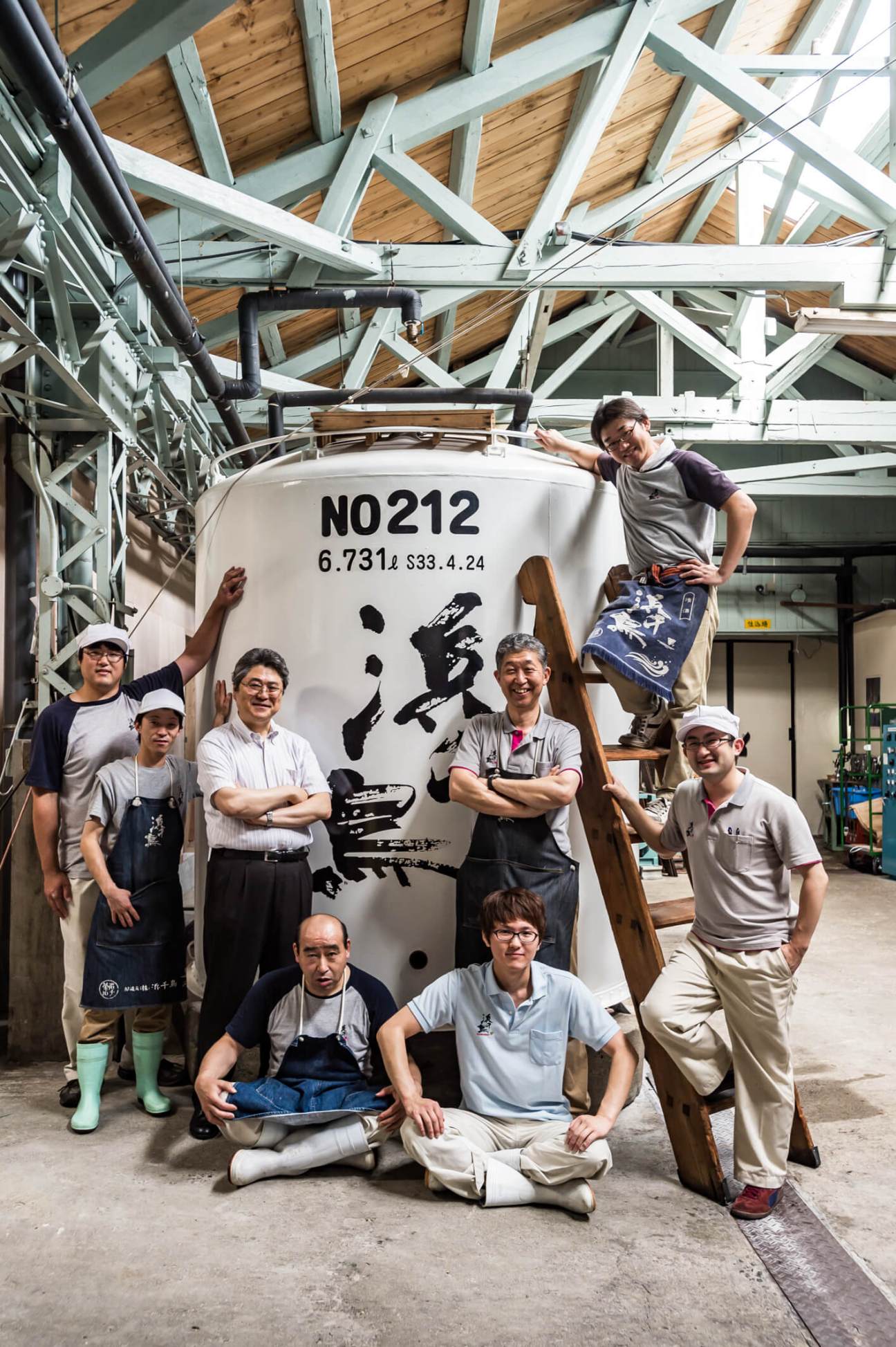 Group of saké makers posing in front of a large white fermentation tank labeled N0212 in a brewery workshop.
