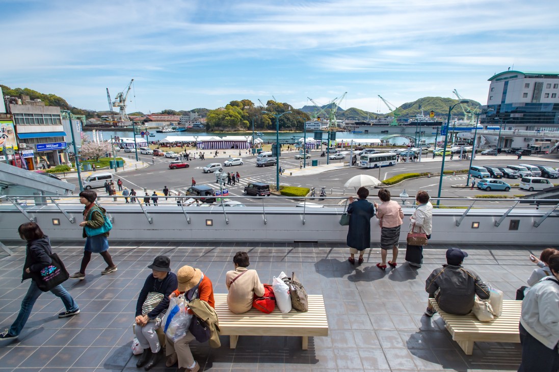 People enjoy an elevated terrace view overlooking Onomichi's waterfront, port facilities, and surrounding hills.