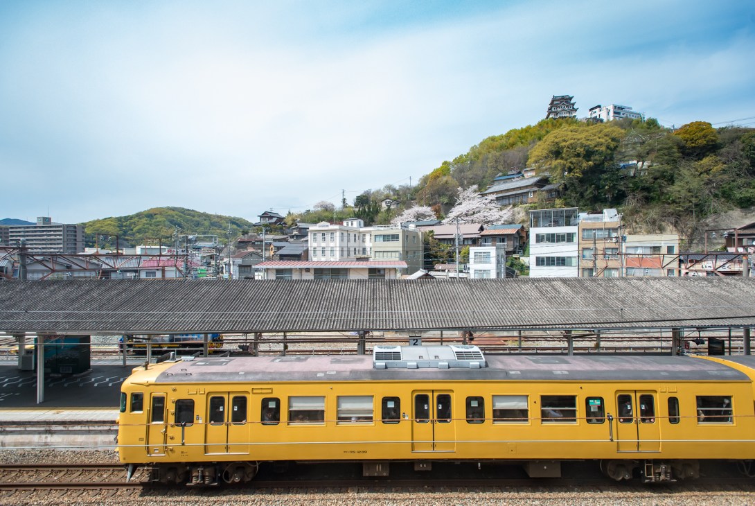 Yellow train at Onomichi Station platform with hillside town and traditional buildings in background.