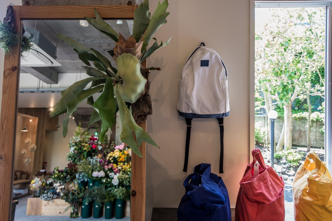 Sunshine + Cloud storefront in Hayama displaying colorful backpacks hanging on wall with potted flowers and green foliage visible through windows.
