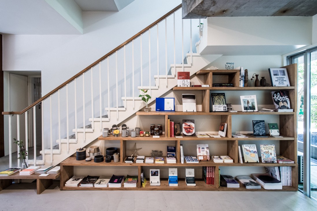 Wooden shelving unit filled with books, magazines, clothing and decorative objects beneath a staircase with white railings.