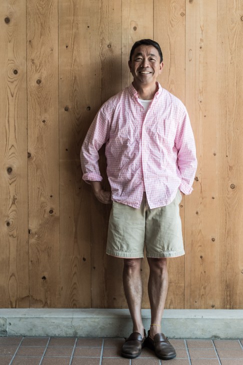 Man in pink checkered shirt and khaki shorts standing against wooden wall backdrop.