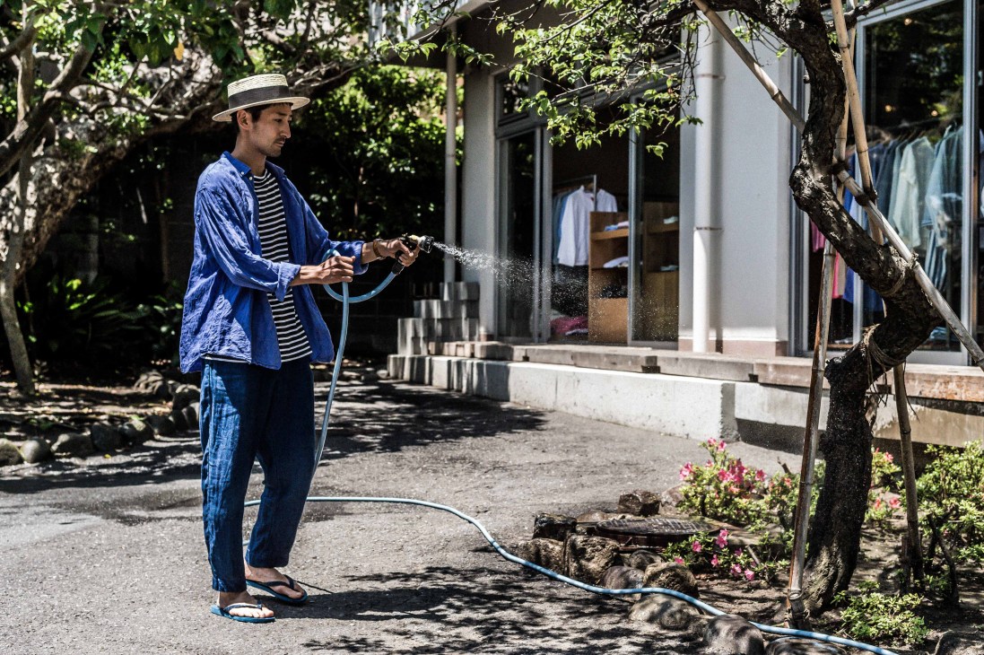 Man in blue shirt watering plants with hose in front of white building with large windows.