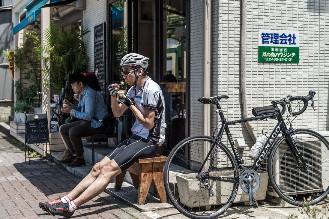 Cyclist in white helmet and cycling gear eating ice cream while sitting on a bench next to parked bicycles on a Japanese street.