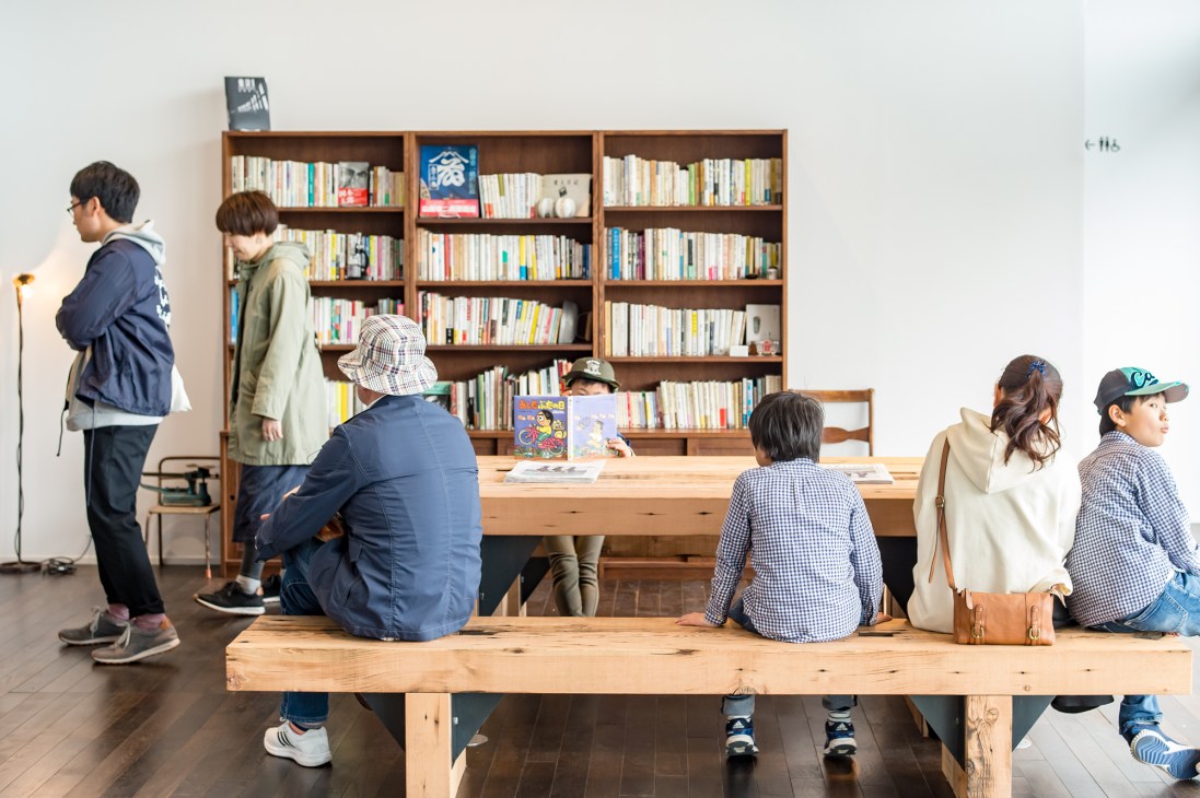 Children and adults browse books in a minimalist lounge area with wooden shelving and natural materials.