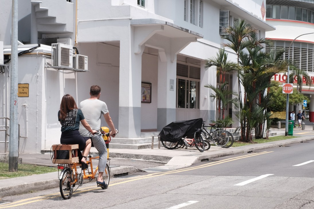 Couple riding a red tandem bicycle on a Singapore street lined with white buildings and palm trees.