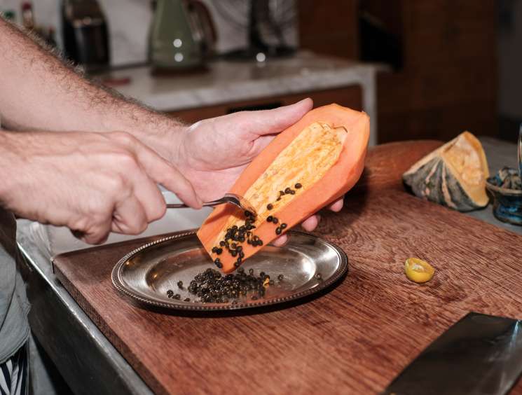 Man's hands halving a papaya over a plate of black seeds at a dining table.