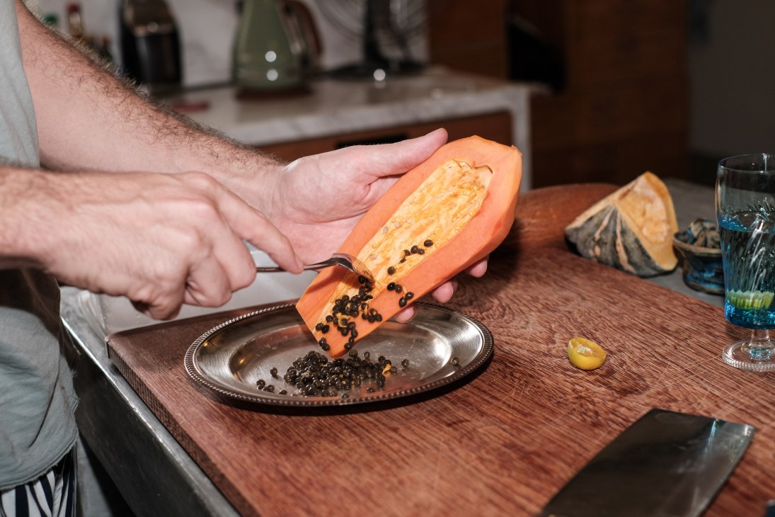 Man's hands halving a papaya over a plate of black seeds at a dining table.