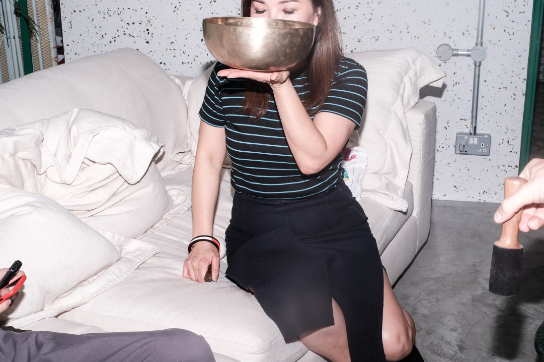 Woman sitting on a bed holding a large brass singing bowl over her face.