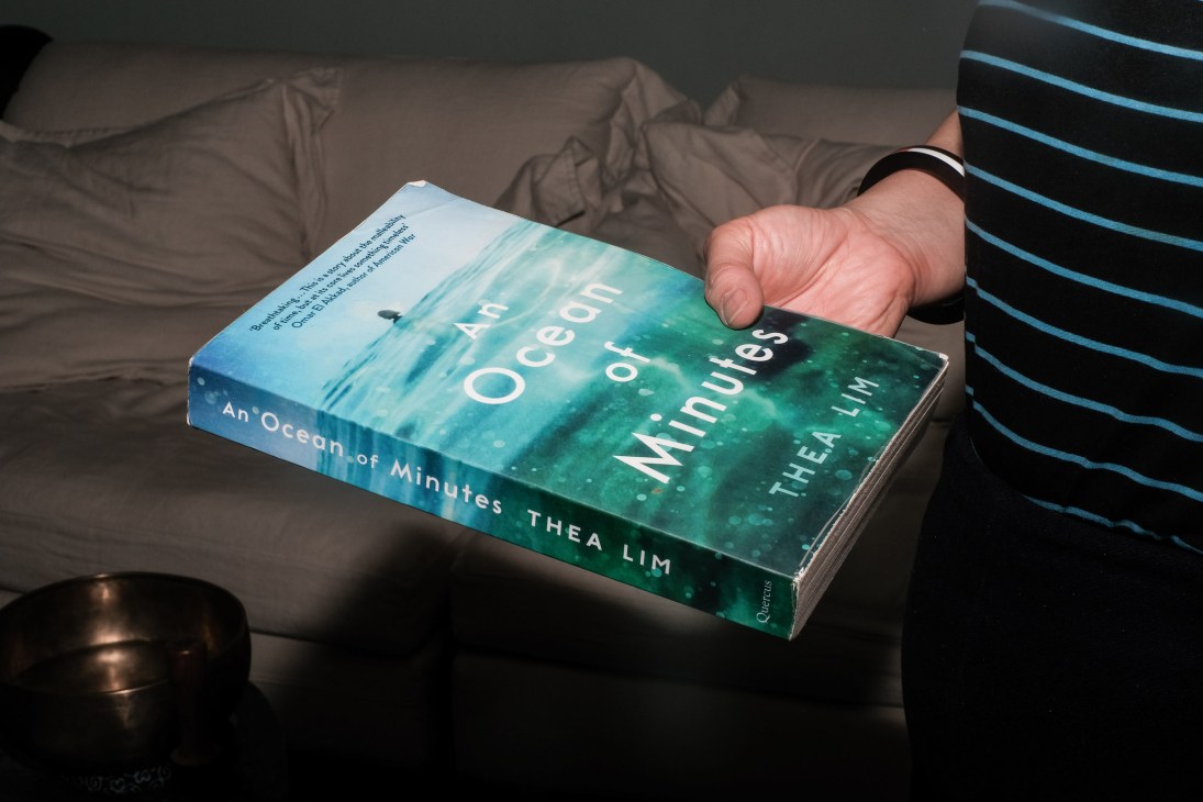 Hand holding the book "An Ocean of Minutes" by Thea Lim with turquoise ocean cover design