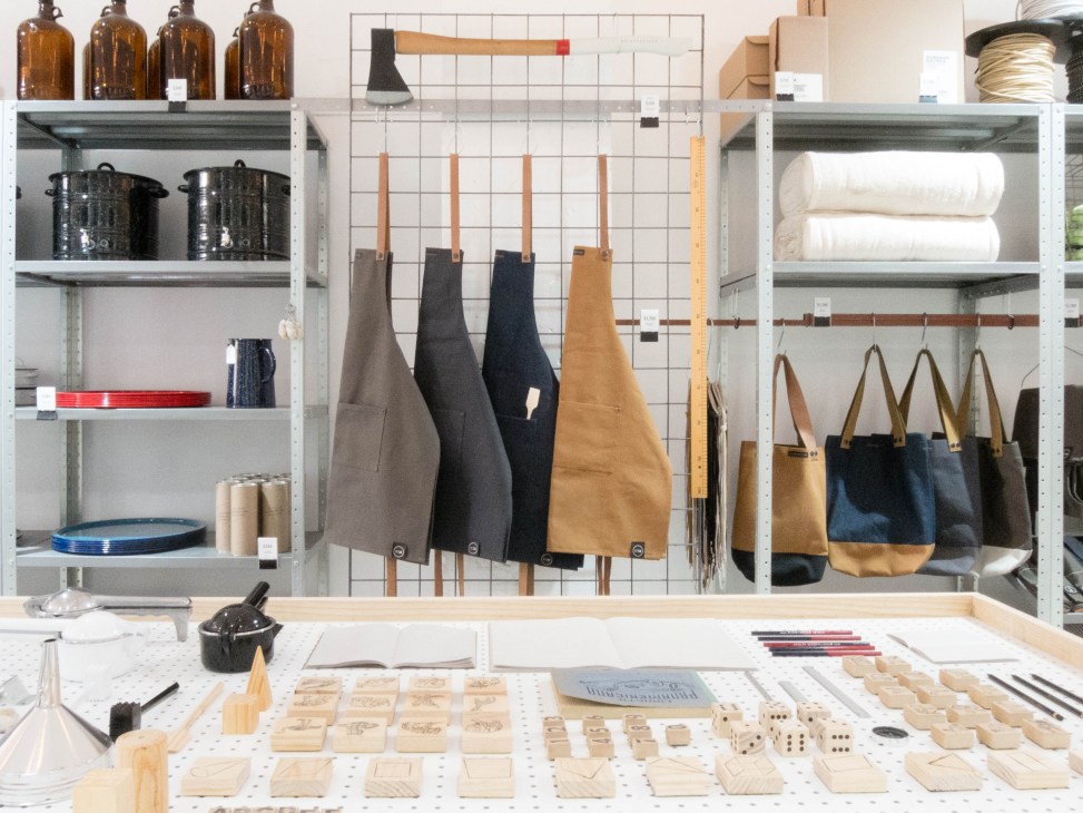 Interior of Utilitario Mexicano shop displaying shelves with cookware, apothecary bottles, rolled towels, canvas tote bags, and wooden craft samples on pegboard wall.