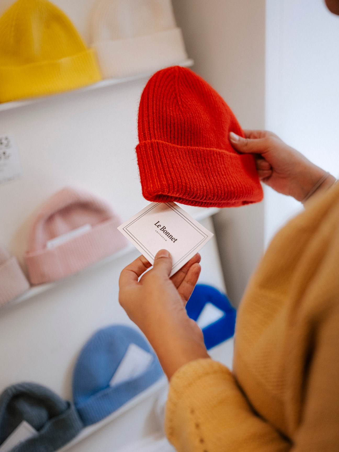 Child holding red knit beanie with gift tag and colorful hats in background