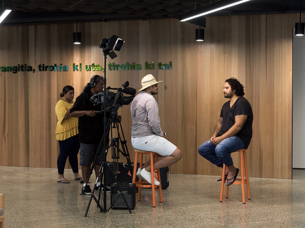 Film crew interviewing Maori musician Troy Kingi in a studio with wooden walls and Maori text signage.