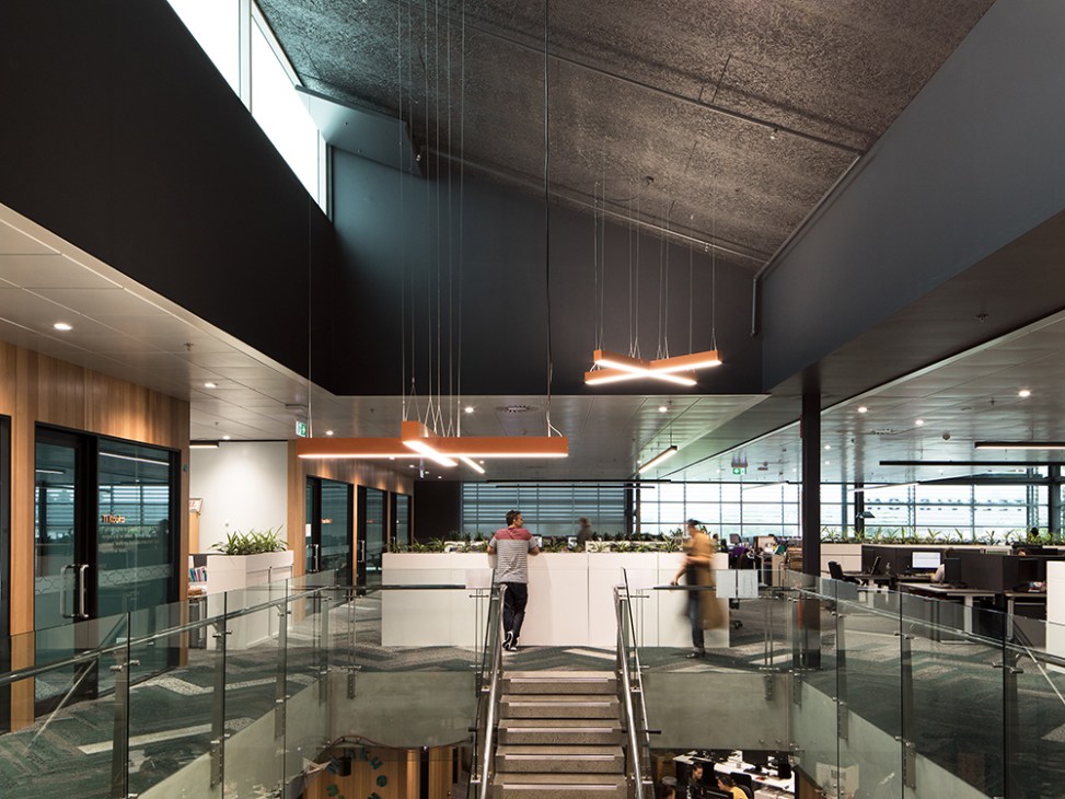 Modern open-plan office space with high ceilings, orange pendant lights, glass railings, and natural daylight from large windows.