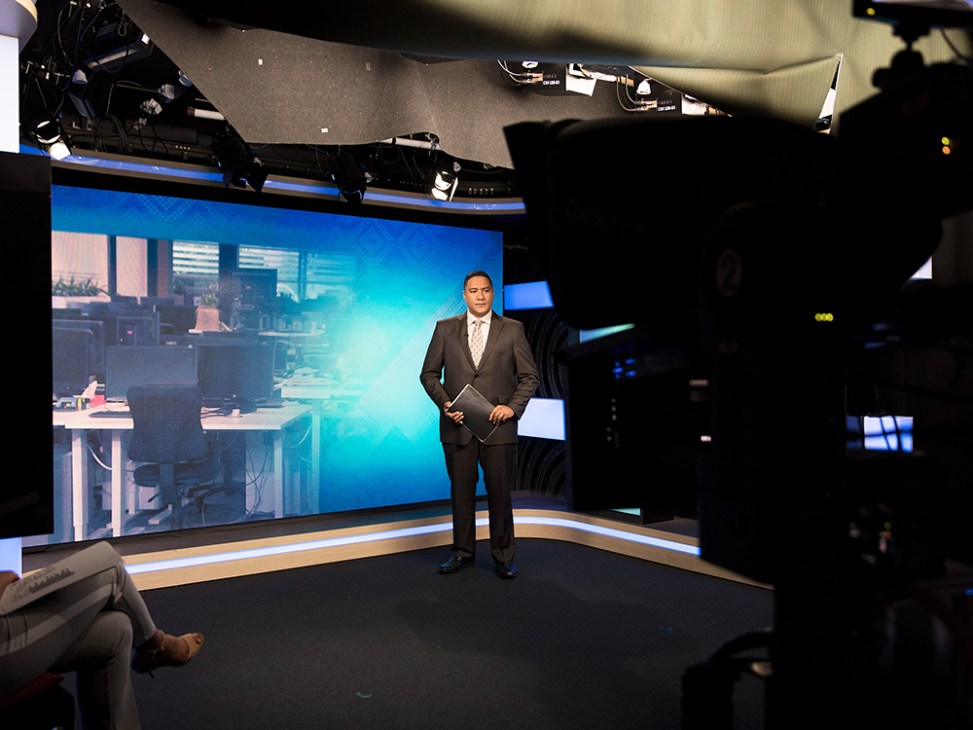Te Kaea presenter Piripi Taylor stands on a modern television studio set with blue lighting and a large screen behind him.