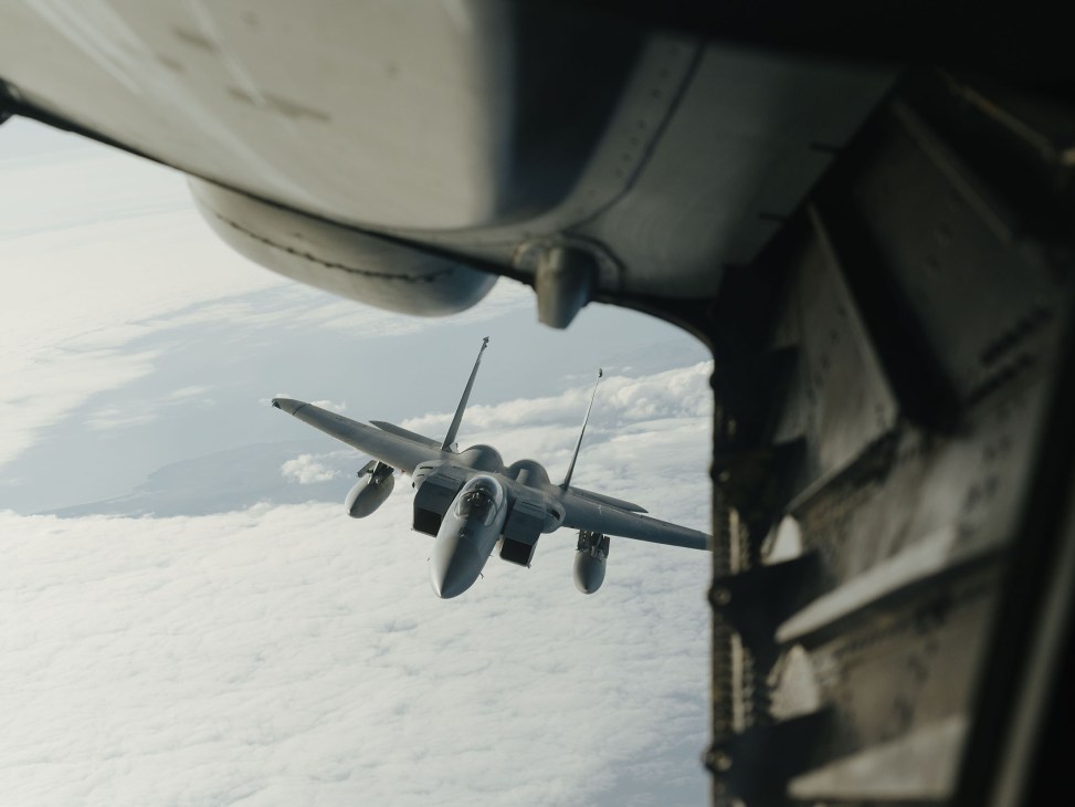 An F-15C Eagle fighter jet approaches an aerial refuelling boom from a tanker aircraft in flight above clouds.