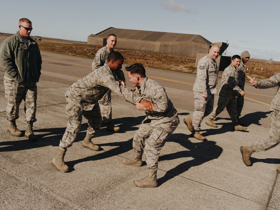 US Air Force personnel in combat fatigues stand together on an airfield with a concrete hangar visible in the background at Keflavík air base.