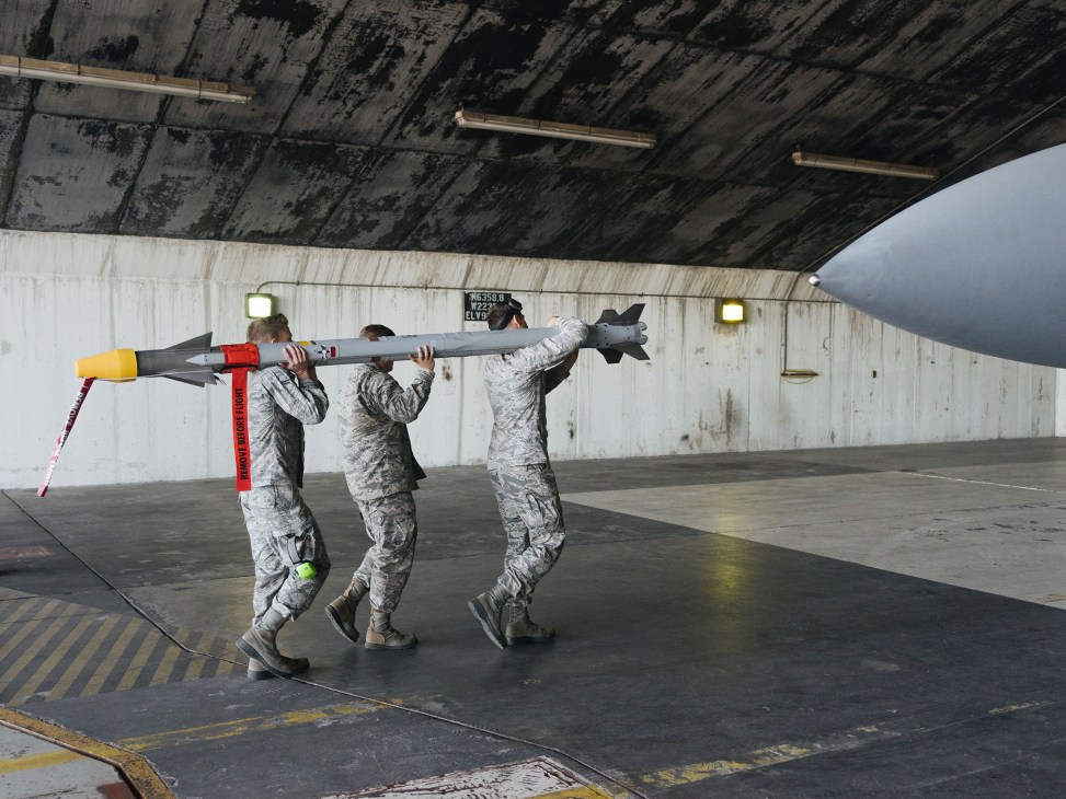 Three US Air Force personnel in camouflage uniforms carry a live missile under an aircraft hangar.