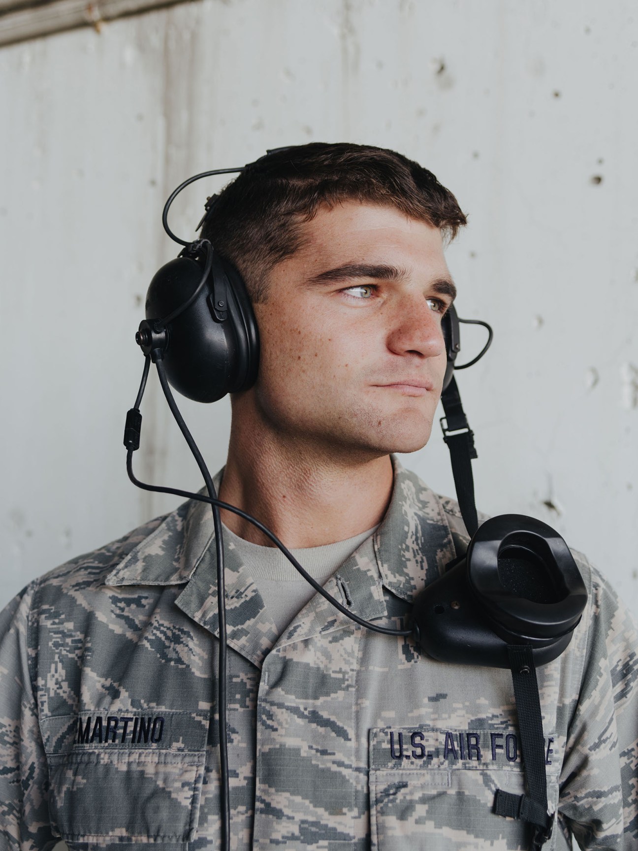 US Air Force airman wearing digital camouflage uniform and black headphones with microphone against white wall.