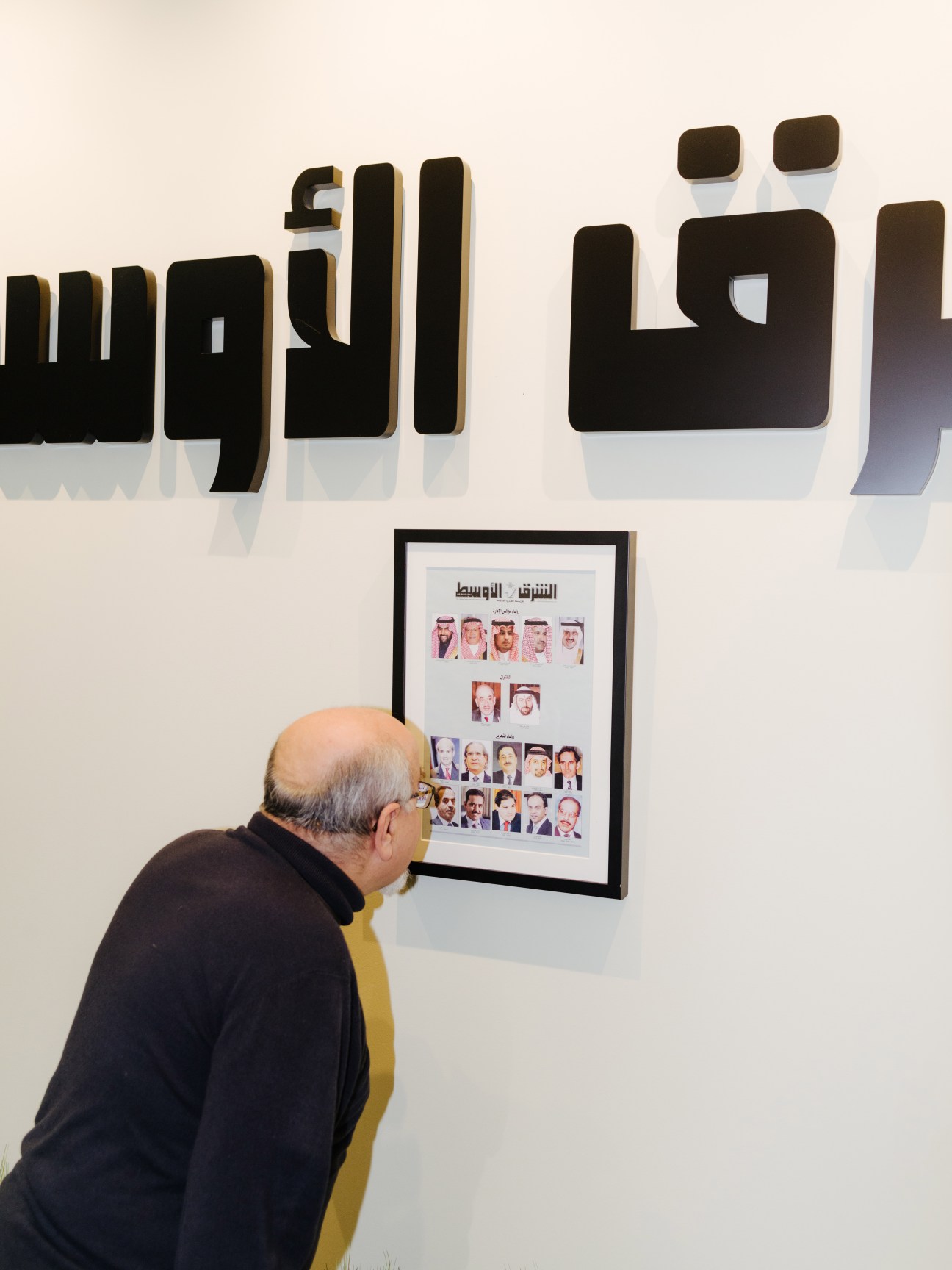 Eyad Abu Shakra viewing a framed display of Asharq Alawsat founders and former editors on an office wall.