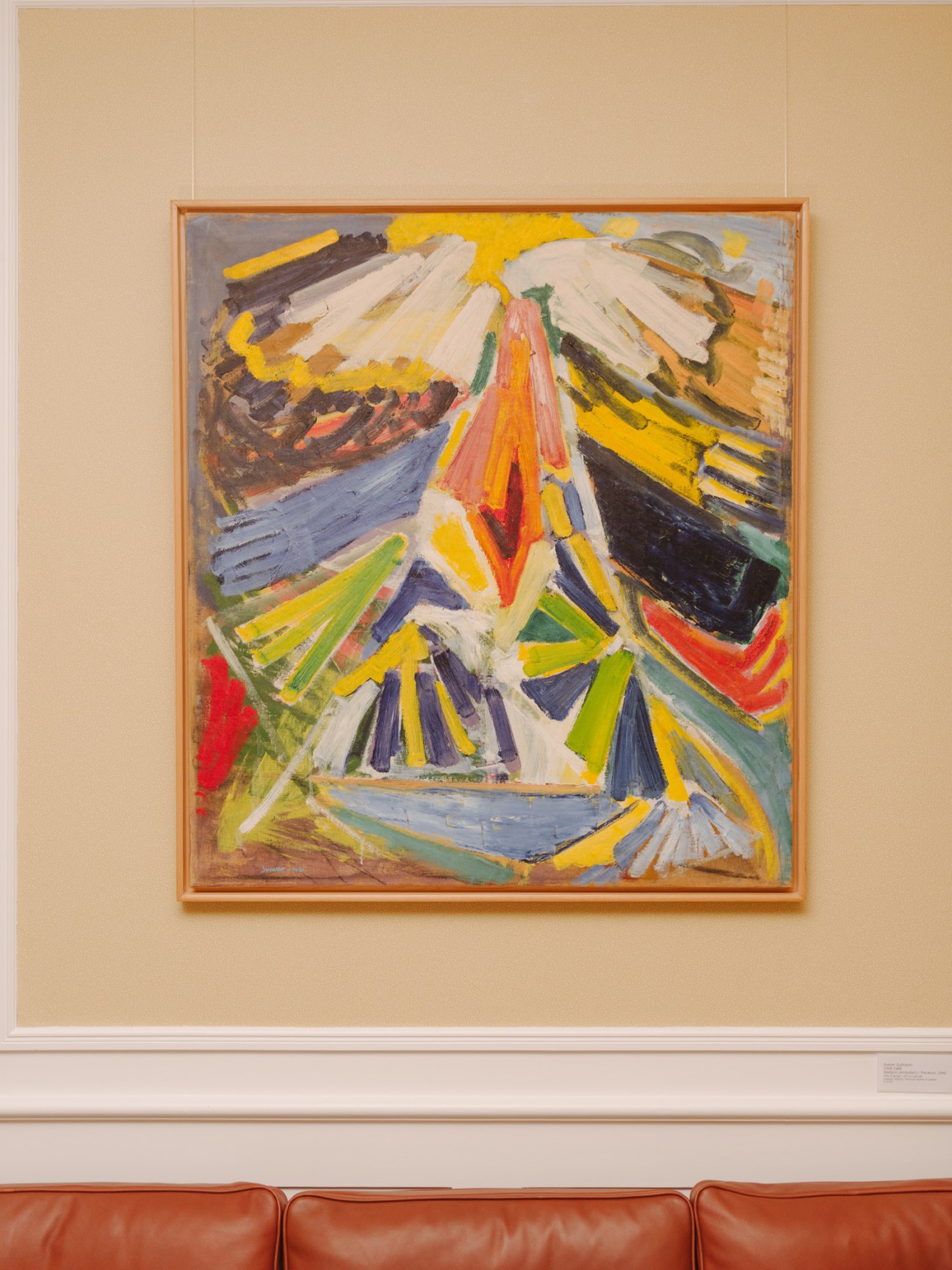 Abstract expressionist painting with bold strokes in yellow, white, red, green, and blue on a cream-colored wall.