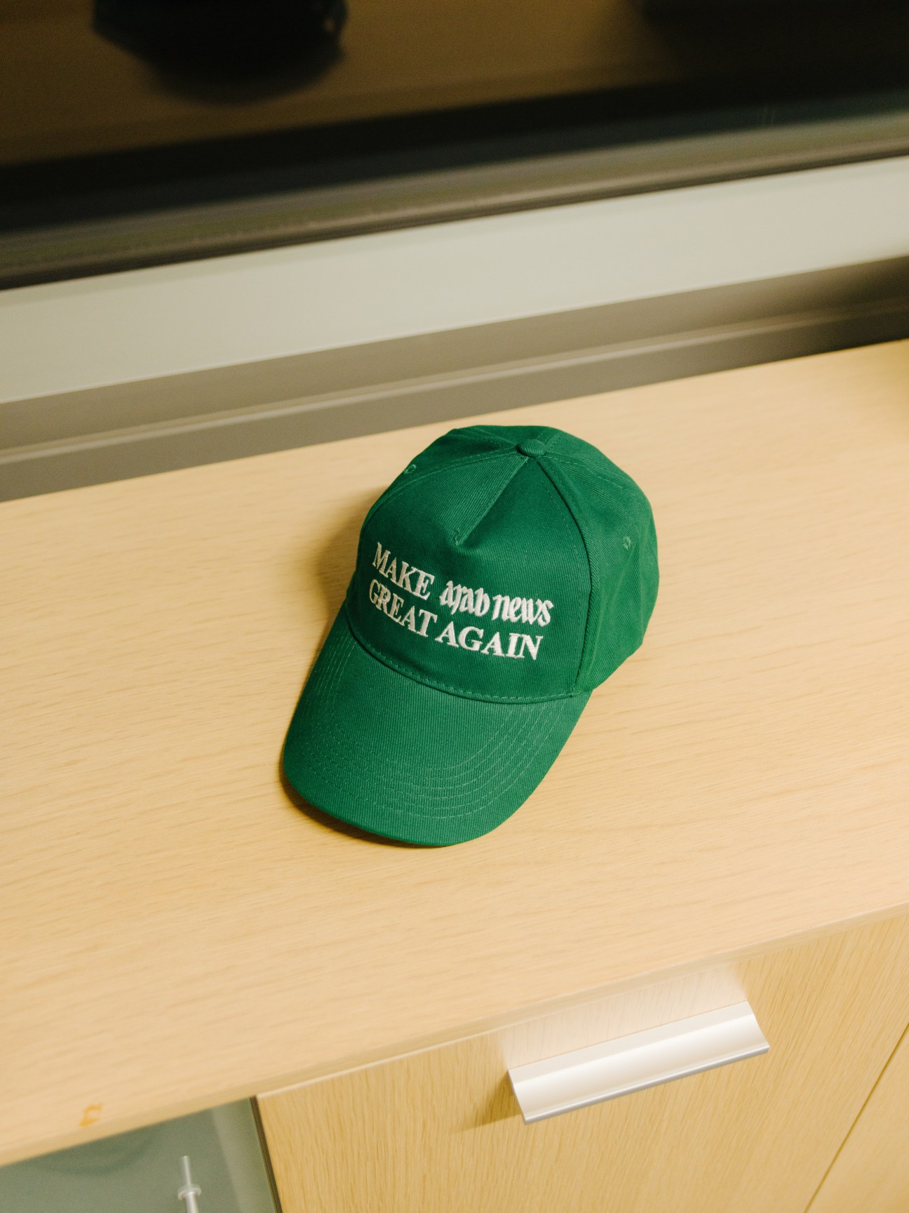 Green baseball cap with 'Make Arab News Great Again' embroidered text on wooden desk