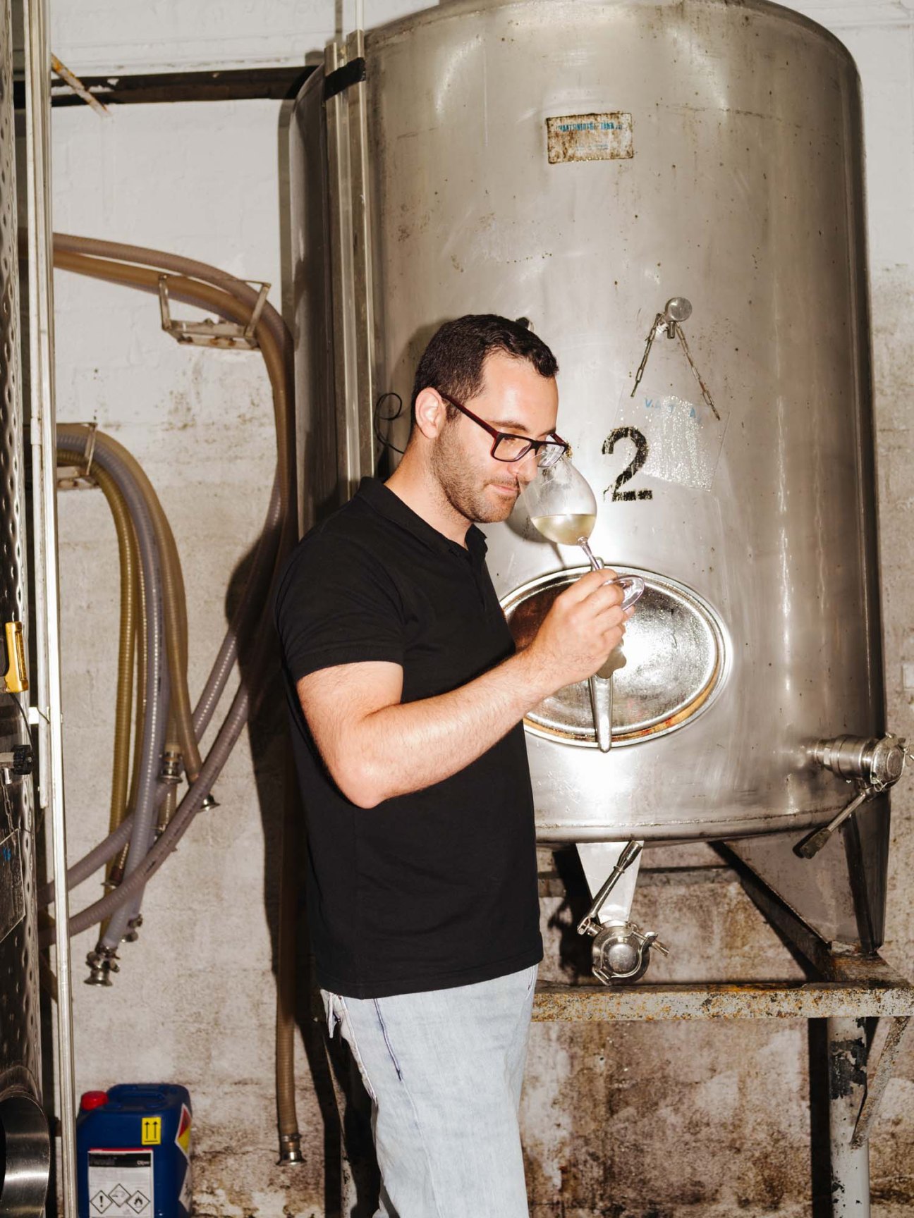 Alexandros Doukas, winemaker at Gentilini, sampling wine from a glass hydrometer against a large metal fermentation tank.