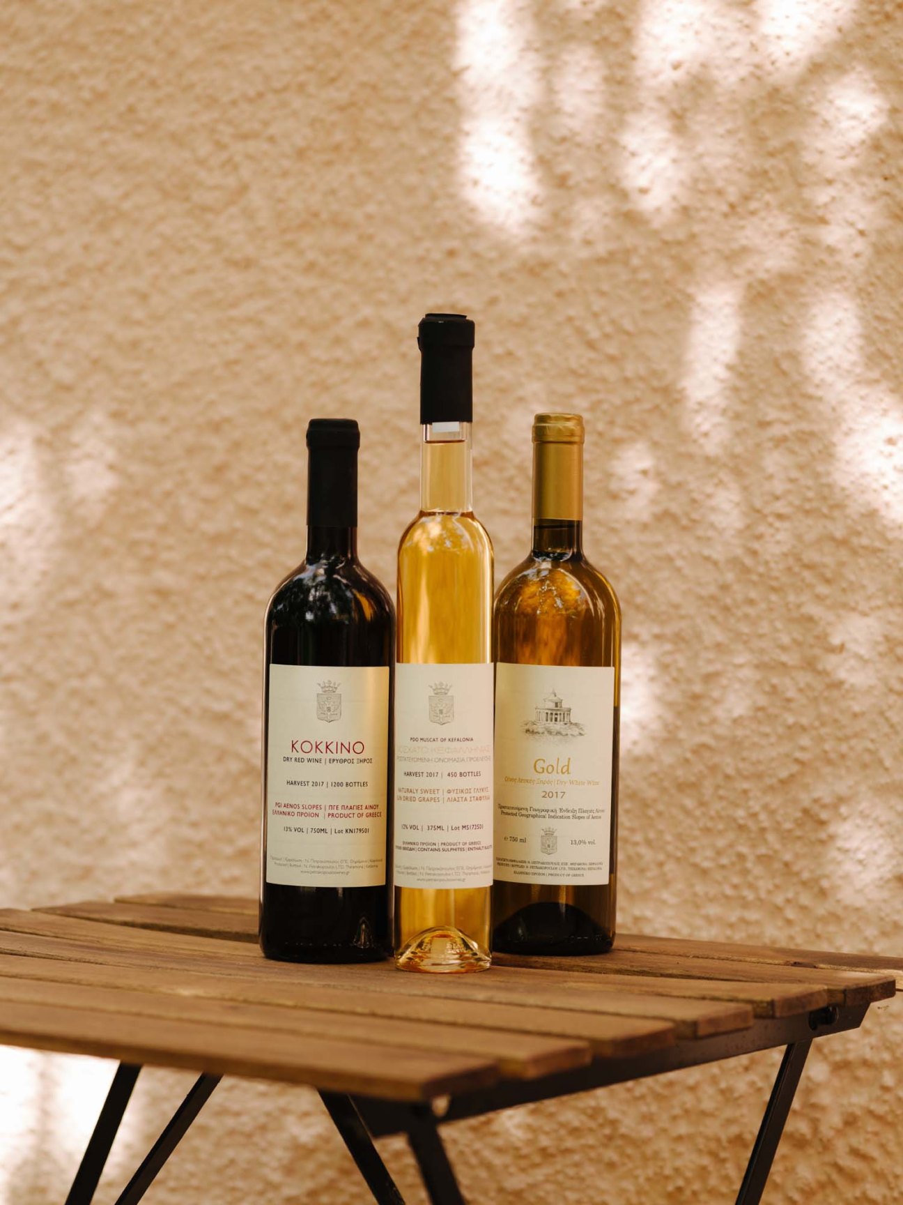 Three wine bottles from Petrakopoulos winery displayed on a wooden table against a textured beige wall.